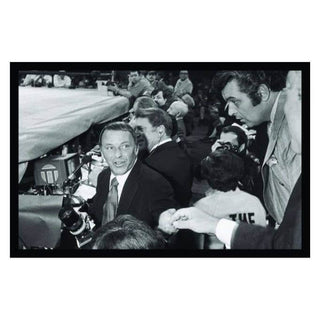 Black and white photo of a smiling man at a boxing event surrounded by celebrities and photographers.