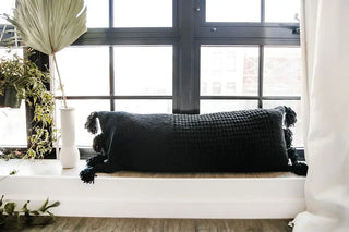 Braided pom pom lumbar pillow in black on a sunny windowsill, featuring a removable cover and memory foam filling.