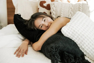 Woman relaxing on a bed with a braided pom pom lumbar pillow, showcasing comfort and style in home decor.