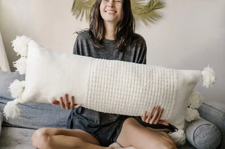 Braided Pom Pom Lumbar Pillow with memory foam and removable cover, held by a smiling woman on a sofa.