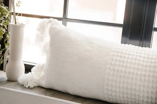 Braided pom pom lumbar pillow with textured cover, styled in a bright, airy room near a window.