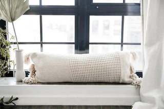 Braided pom pom lumbar pillow on a windowsill, featuring a textured cover and decorative tassels, perfect for stylish decor.