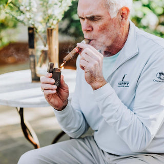 Man igniting a cigar with a custom Colibri Julius lighter in a serene outdoor setting.