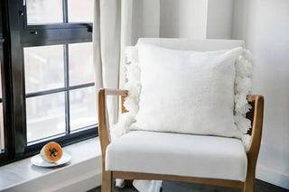 Pom Pom Throw Pillow in soft microfiber with fuzzy pom poms, placed on a cozy chair by a window.