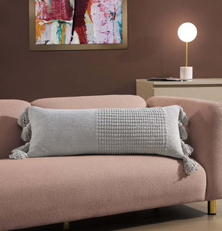 Braided pom pom lumbar pillow in light gray on a pink couch, featuring a textured cover and decorative tassels.