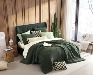 Cozy green bedroom with a plush bed featuring various cushions, a knitted throw, and a large window letting in natural light.