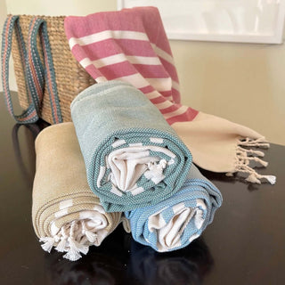 Rolled beach towels in beige, green, and blue colors next to a woven bag and striped towel on a dark table.
