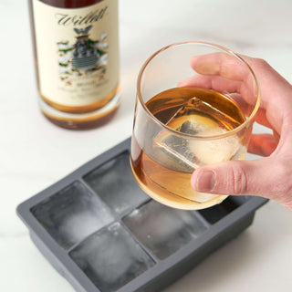 Hand holding a glass of whiskey with oversized ice cubes from a flexible silicone ice tray beside a whiskey bottle.
