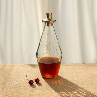 Handcrafted pebbled glass decanter with hammered aluminum stopper, filled with rich liquid, on a textured surface.