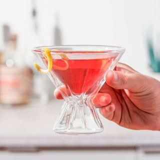 A hand holding a double-walled martini glass filled with a red cocktail and garnished with a lemon twist.