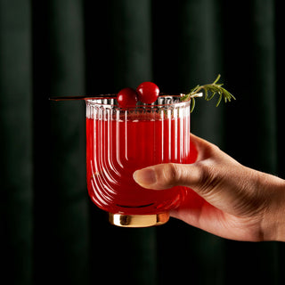 Elegant hand holding a Gatsby tumbler filled with a red cocktail, garnished with cherries and herbs.