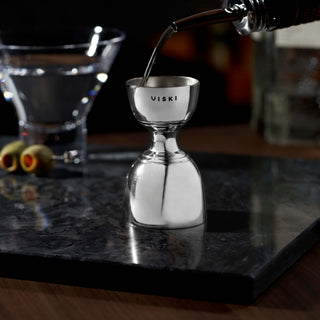 Harrison Canterbury Jigger pouring liquor with a cocktail and olives in the background on a stylish bar top.