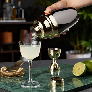 A person pouring a cocktail from a black and gold heavyweight shaker into a glass garnished with lime.