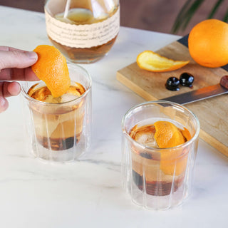 Double-walled rocks glasses set of 2 with cocktails, garnished with orange peel, on marble countertop.